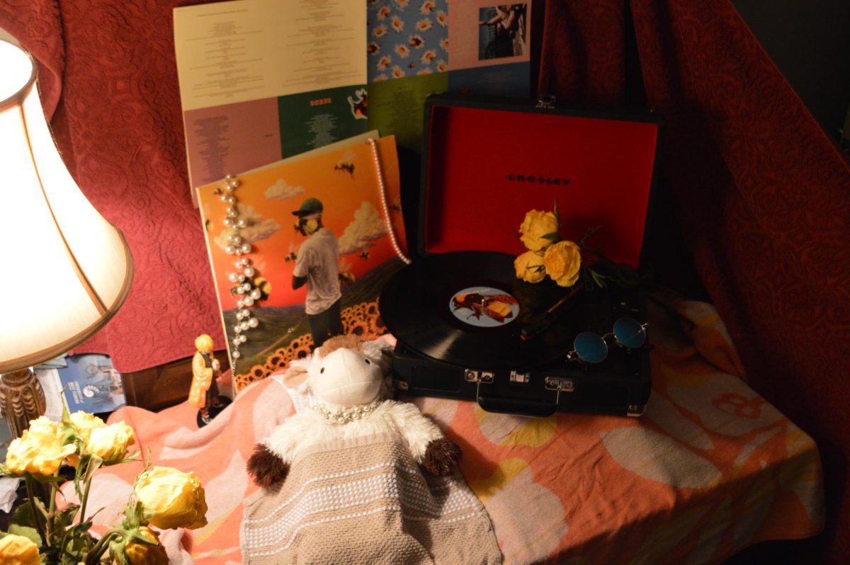 This photograph captures a stylized still life centered around the album “Flower Boy” and its cozy and intimate feelings along with a Crosley suitcase turntable and a stuffed animal. I used different boxes that I had around my house to create levels for the record player and the album itself, while also creating a level for the stuffed cow's pillow to create more of a higher perspective for the cow. For the light source, I used a small and relatively dim lamp to create more shadows and cause a warm, dreamy feeling in the picture. I styled the subjects by applying different jewelry to hang off the album to give it a shinier and delicate look. I also put a small piece of jewelry on the stuffed animal to draw some attention to it as well. Along with that I put a record from the album onto the Crosley, plus some yellow flowers and some blue tinted sunglasses so it wasn’t as empty. The background is a dark red with slight patterning and the base is covered in a warm orange and tan mix.
